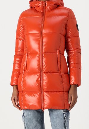 Lang orange dundunjakke med hætte, frontlynlås, sidelommer og en sort cirkulær patch på venstre ærme, båret med lyseblå cargobukser.