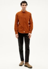 Rostfarbendes, langärmliges Hemd mit Kragen und einem kleinen Logo-Patch, kombiniert mit schwarzen Hosen und braunen ankle boots.