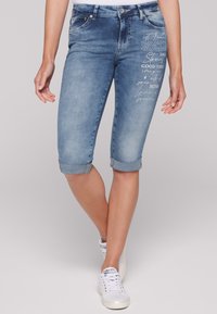 Bermudas de mezclilla en azul claro con puños enrollados, que presentan texto blanco y pequeños acentos decorativos en el muslo izquierdo.
