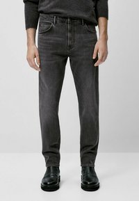 Man i mörkgrå slim-fit jeans, svarta läderstövlar och en mörkgrå tröja, stående mot en enfärgad ljus bakgrund.
