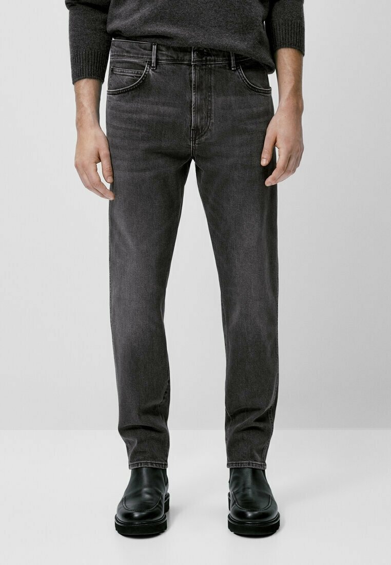 Man i mörkgrå slim-fit jeans, svarta läderstövlar och en mörkgrå tröja, stående mot en enfärgad ljus bakgrund.