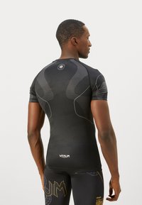 Homme athlétique de dos portant un t-shirt de compression noir avec des panneaux à motifs et un legging noir avec des designs jaunes et gris.