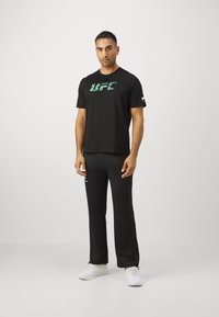 Črna majica s kratkimi rokavi z zelenim logotipom "UFC", v kombinaciji s črnimi hlačami. Oboje je izdelano iz mehke tkanine; čevlji so beli z teksturirano obdelavo.