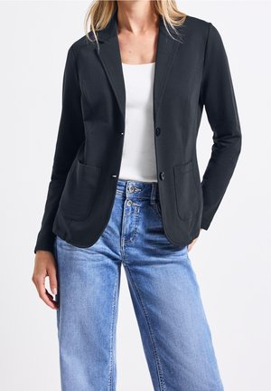 Kvinde iført sort blazer over hvid top og lyseblå højtaljede jeans, stående med den ene hånd i lommen mod en hvid baggrund.