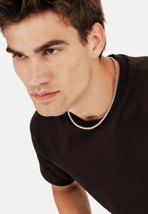 Camisa negra de manga corta con cuello redondo, presentada con un collar de cadena plateada. El fondo es blanco liso, lo que resalta la camisa y el collar.