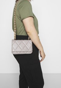 Persona che indossa una camicia a maniche corte verde oliva e pantaloni neri, con una piccola borsa trapuntata grigio chiaro con tracolla dorata a spalla.