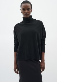 Maglione nero a collo alto con una vestibilità comoda, polsini e orlo a coste. Texture morbida, maniche lunghe e design minimalista, indossato con una gonna nera.