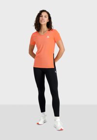 Camiseta atlética de manga corta en color naranja con un panel lateral negro, combinada con leggins negros. Presenta un sutil logo en el pecho y acentos en blanco.