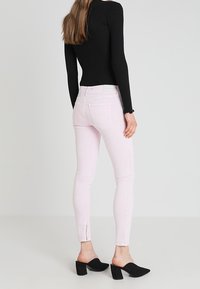 Vaqueros skinny de color rosa claro con una textura suave, con cintura de altura media y cremalleras en los tobillos, combinados con una blusa ajustada negra y mules negros.
