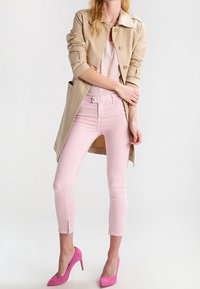 Mujer con un abrigo trench beige, pantalones rosa claro con cremalleras en los tobillos, y tacones altos rosa brillante, de pie contra un fondo blanco.