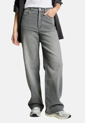 Person wearing loose gray jeans with one hand in pocket and white sneakers, standing against a plain background.