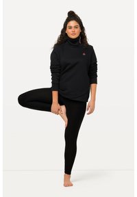 Pull noir à col montant avec des poignets repliés, associé à des leggings noirs texturés. Le mannequin se tient sur une jambe, mettant en valeur le motif et l'ajustement des leggings.