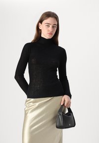 Maglione nero a coste con collo alto, aderente, con maniche lunghe. Il modello indossa una gonna midi dorata lucida e tiene una piccola borsa nera.