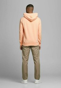 Pfirsichfarbene Kapuze Sweatshirt mit weicher Textur, kombiniert mit hellbraunen Hosen und weißen Sneakers, von hinten betrachtet.