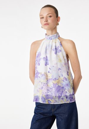 Kvinde iført hvid ærmeløs halterneck med lilla blomsterprint og høj krave, kombineret med mørkeblå jeans, stående mod en hvid baggrund.