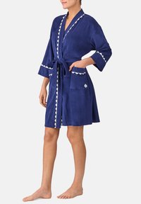 Donna che indossa un abito blu navy al ginocchio con bordo bianco scallopato, legato in vita, con tasche frontali, in piedi a piedi nudi su uno sfondo bianco.