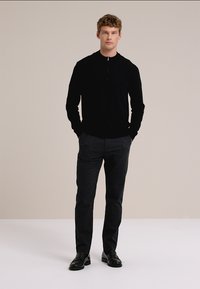 Maglione nero con zip, pantaloni aderenti grigi e scarpe nere. Il modello è in piedi diritto con le mani nelle tasche su uno sfondo neutro.