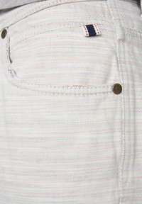 Tissu en denim blanc avec des rayures horizontales multicolores. Comprend un bouton en métal et une petite étiquette bleu marine et blanche sur la poche.
