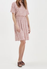 Midi šaty s květinovým vzorem v růžové a bílé látce, mají krátké rukávy, elastický pas a sahají těsně nad kolena, nošeny s tmavými sandály.