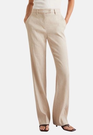 Pantaloni beige a gamba larga con piega frontale, indossati con top bianco senza maniche e sandali neri con tacco e punta quadrata. Mani nelle tasche.