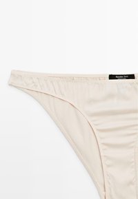 Light beige thong underwear made of smooth fabric. Features a simple design with minimalistic stitching and a small logo tag.