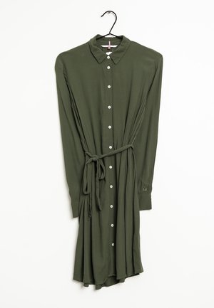 Langærmet olivengrøn skjortekjole med knapper, krave og taljebånd, hængende på en sort bøjle mod en hvid baggrund.