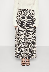 Kvinna som bär vidbeniga plisserade byxor med zebramönster i svart och vitt, en krämfärgad stickad tröja och svarta sandaler med klack och remmar.