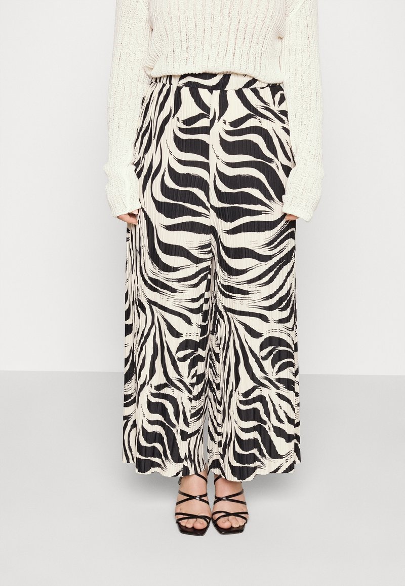 Kvinna som bär vidbeniga plisserade byxor med zebramönster i svart och vitt, en krämfärgad stickad tröja och svarta sandaler med klack och remmar.