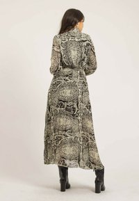 Robe maxi en tissu transparent à motifs avec des designs noirs et beiges. Manches longues, taille ceinturée et silhouette fluide. Bottes noires à la cheville.
