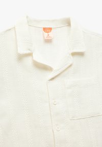 Weißes Baumwollbabyshirt mit Kragen, Brusttasche, strukturierten Streifen und Knopfschluss. Pflegeetikett ist sichtbar.