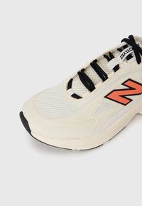Pantof de sport bej cu șireturi negre, logo-ul portocaliu "N", superior din plasă și vârf din cauciuc negru pe un fundal alb simplu.