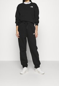 Schwarzes Sweatshirt- und Jogginghosen-Set aus weichem Stoff, mit einem geprägten Logo auf der Brust und am Bein. Kombiniert mit weißen Sneakers.