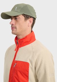 Veste en polaire beige avec un panneau de poitrine orange, col haut et fermeture éclair. Casquette vert olive avec des côtés perforés et logo.