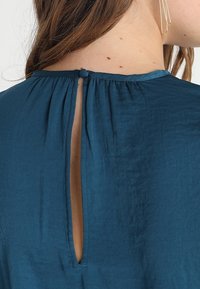 Teal blus med plisserad baksida, vertikalt utskärning och knäppning i nacken. Slät textur och lättviktig tyg.