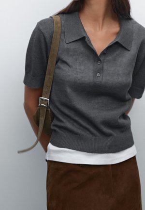 Woman wearing a dark gray short-sleeve polo shirt, white undershirt, brown skirt, and carrying an olive green shoulder bag.