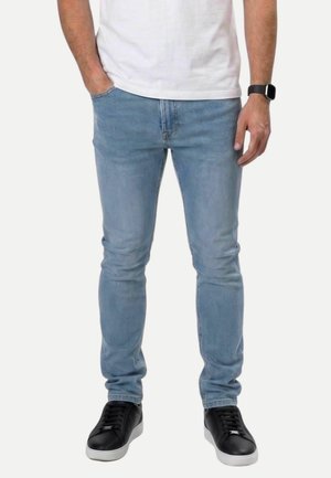 Homme portant un jean slim bleu clair, une chemise blanche, des baskets noires à semelles blanches, et une montre connectée noire au poignet gauche. Mains dans les poches.