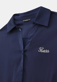 Granatowa koszula z guzikami, z kołnierzykiem, gładką tkaniną i srebrnym logo „Guess” z cyrkoniami po lewej stronie.