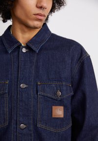 Denim jacket in dark blue with yellow stitching, featuring two chest pockets, snap buttons, and a leather brand tag on the left pocket.