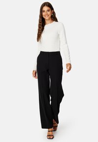 Witte gestructureerde langemouwen top gecombineerd met zwarte wijde broek en zwarte bandjes sandalen, wat een strak, eigentijds uiterlijk laat zien.