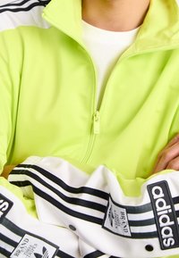 Neon green zip-up jacket with a high collar; white cuffs featuring black stripes and branding details; smooth, lightweight material.