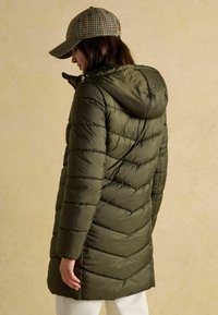 Olive zelená puffa bunda má vertikálne a diagonálne prešitie, kapucňu a zips vpredu, kombinovaná s kockovanou čiapkou.