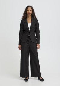 Blazer noir cintré à un bouton, associé à un pantalon noir à jambes larges et des chaussures plates noires, sur un fond blanc.