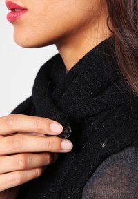 Svart stickad väst med hög krage, som har en framträdande rund svart knapp nära halslinningen, texturerad tyg och en tajt passform.