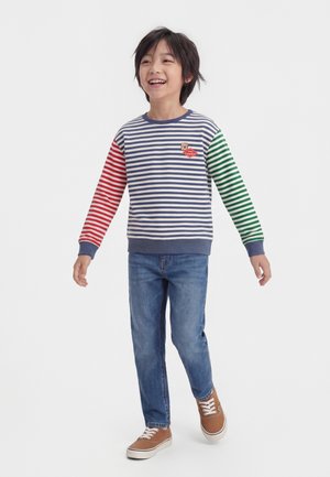 Smilende barn iført stribet langærmet skjorte med røde og grønne ærmer, blå jeans og brune sneakers, står med armene let ud til siden.