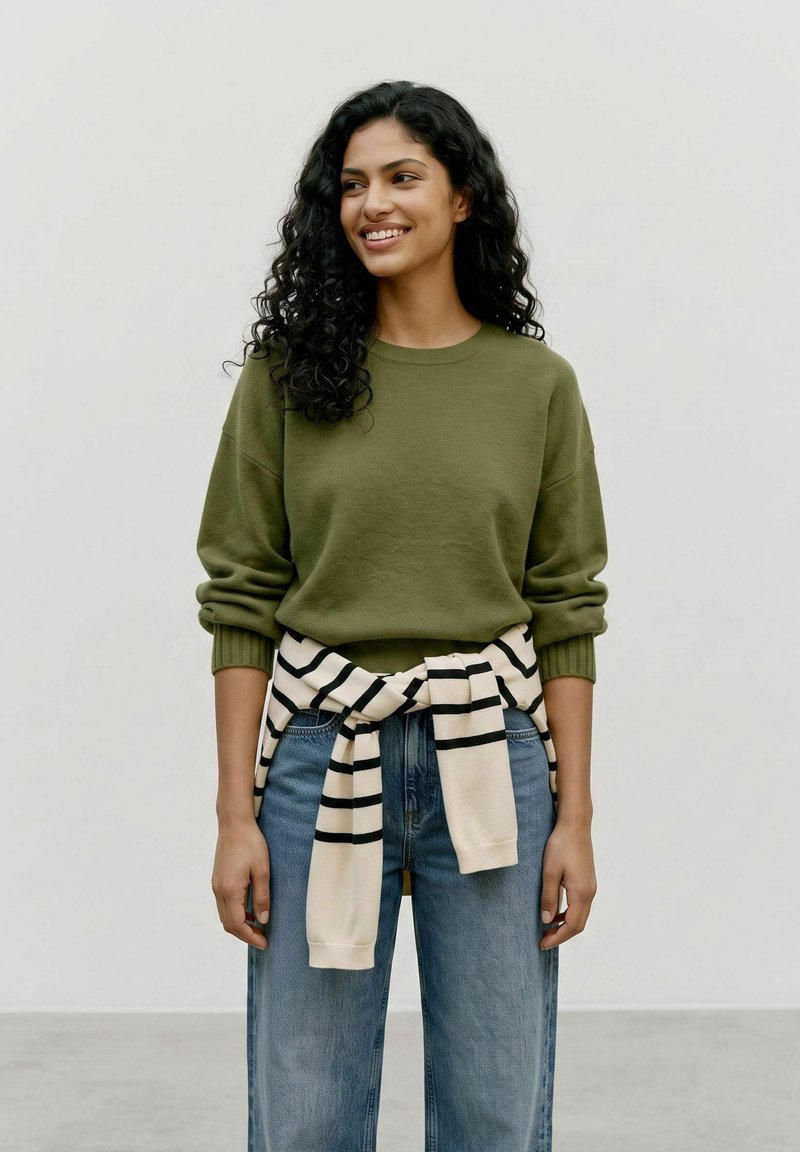 Jeune femme aux cheveux noirs bouclés portant un pull vert olive et un jean bleu avec un pull rayé noué autour de la taille, souriante.