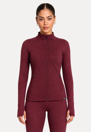 Femme portant une veste zippée ajustée couleur bordeaux et des leggings assortis avec un imprimé léopard subtil et des trous pour les pouces sur les manches.