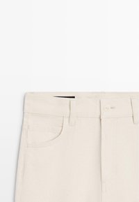 Pantalon en denim beige à jambe droite, doté d'une fermeture par bouton, d'une poche avant et de passants pour ceinture. Texture lisse avec des détails minimes.