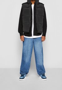 Gilet imbottito nero, camicia nera con maniche lunghe e bottoni, t-shirt bianca sotto, e jeans in denim blu larghi con sneakers blu.