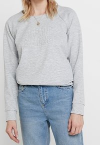 Sudadera gris con un logo "ARMANI EXCHANGE" texturizado y en relieve. Presenta mangas raglán y un corte relajado. Combinada con jeans de color azul claro.