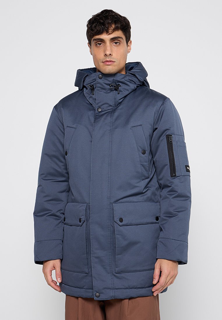 Navyblauwe gewatteerde jas met capuchon, voorzakken en een met rits afsluitbare mouwzak. Bevat drukknoopsluitingen en verstelbare trekkoorden.